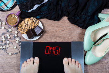 Girl checks her weight on digital scales. Sign omg! on balance surrounded by cup of coffee, sweets and female clothes and shoes. Concept of unhealthy eating and its consequences.