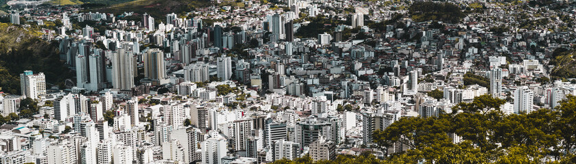 Panoramic shot of an urban landscape from high above of Juiz de Fora town in Minas Gerais state of Brazil: multiple multistorey residential and office buildings, favelas, parks, and hills, bright day