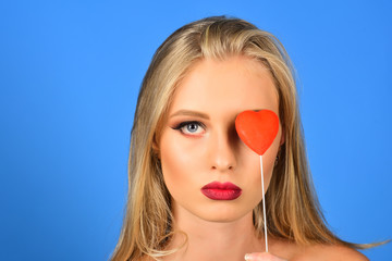 Valentines day love and relationships concept. Beautiful gorgeous woman with glamour bright makeup, red lips, perfect skin holding heart on eye. Isolated on blue background. Copy space for advertising