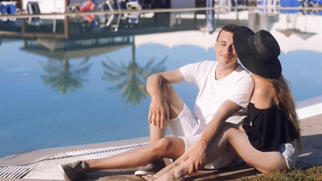 Carefree lovers resting near swimming pool. Girl in sunglasses and hat. Couple on vacation, sunbathing, relaxing. Beackground palms and pool. Vacation, tourism, hooneymoon.