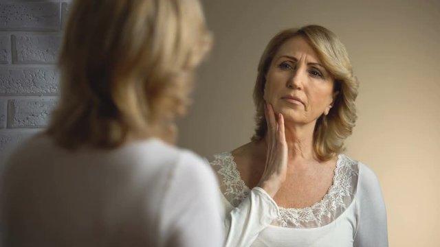 Retired woman sadly looking into mirror, age appearance problem, wrinkles