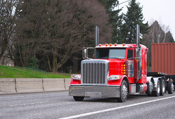 Bright red classic big rig semi truck transporting container on flat bed semi trailer on wide multiline highway