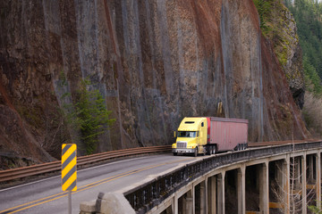 Yellow big rig semi truck carry cargo in bulk semi trailer on winding dangerous road with rock wall and bridge