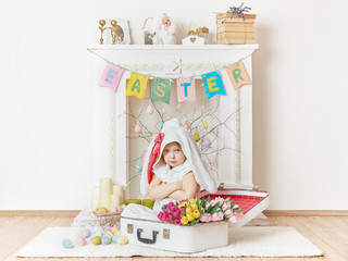 Little girl in white bunny costume sitting in vintage suitcase with her arms folded across her chest; Easter decorated fireplace on background