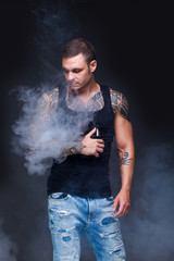 Vaper. The man with a muscular torso with tattoos smoke an electronic cigarette on the dark background