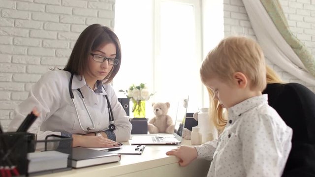 Kind and sweet woman doctor talking with a little boy in a children's clinic. Mom with a little son at a pediatrician, the doctor is very nice talking with the baby. Little boy is afraid of the doctor