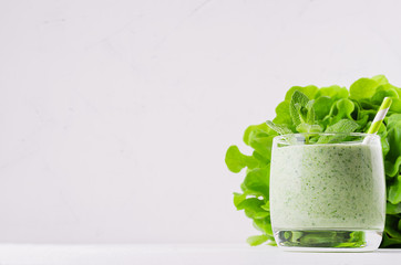Freshly blended green spinach smoothie in glass with straw, mint leaves, sheaf salad. White wooden board background, copy space.