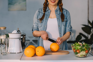 cropped image of smiling pregnant woman cutting oranges at kitchen