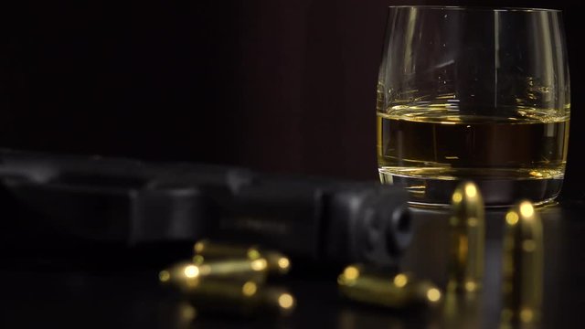 Closeup on a glass of whiskey on a black table - a gun and bullets in the blurry foreground