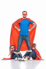 super dad standing with hands on waist and looking at camera while kids hugging his legs isolated...
