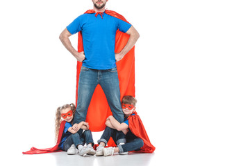 cropped shot of super dad standing with hands on waist while kids hugging his legs isolated on white