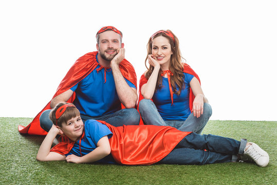happy family of superheroes sitting on lawn and smiling at camera on white