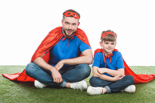 happy father and son in superhero costumes sitting on lawn and smiling at camera on white
