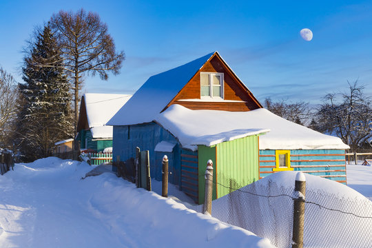 Typical Russian dacha, blue sky with  Moon. Cabins painted with vivid colors look great surrounded with snow
