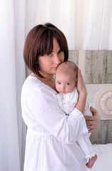 Young mother is holding her little baby. Woman hugs her newborn child. Bit sad look. White light interior