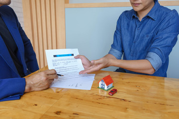 Close up of customer signing a paper document for buying house. Estate agent pointing finger showing where to sign on wooden desk: real estate, home loan and insurance concept