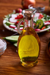 Golden olive oil in transparent glass jar in front of plate with vegetable salad, shallow depth of field