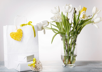 Shopping bag, yellow heart and ribbon, gift and white bouquet tulips bouquet in a vase on white background.