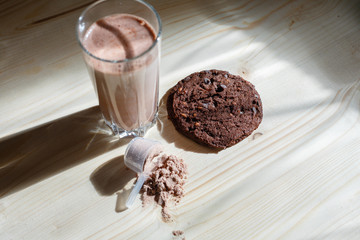 Mixed milk chocolate with whey protein shake in glass cup, scoop with protein and chocolate cookie on wooden table. Dietary snack for bodybuilding. Protein casein, geiner, hydrolysate after training