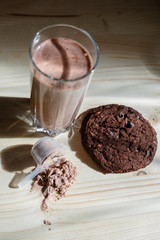 Mixed milk chocolate with whey protein shake in glass cup, scoop with protein and chocolate cookie on wooden table. Dietary snack for bodybuilding. Protein casein, geiner, hydrolysate after training