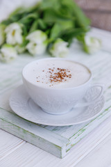 Coffee cappuccino with cinnamon in a white cup and white tulips on a light wooden background