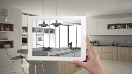 Hand holding tablet showing kitchen sketch or drawing. Real finished modern kitchen with island and stools in the background, architecture interior design presentation