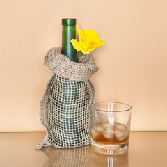 Glass of whiskey with ice and a bottle wrapped in decorative canvas bag with daffodil.