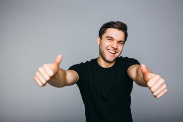 Fototapeta premium Happy successful confident man in black t-shirt showing thumbs up . Handsome young man gesturing and keeping mouth open while standing against grey background