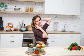 Attractive blonde girl smiles and looks at the camera. A young woman sits at a kitchen table with vegetables and fruits in the dining room.