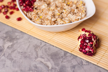 Cooked yummy warm oatmeal and heap of ripe fresh pomegranate seeds in white ceramic bowl near piece of pomegranate on clean bamboo rug on worn gray scratched concrete with copy space