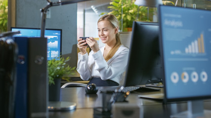 In the Office Attractive Businesswoman Plays Video Games on Her Smartphone. Smiling Beautiful Woman Sitting at Her Desk.