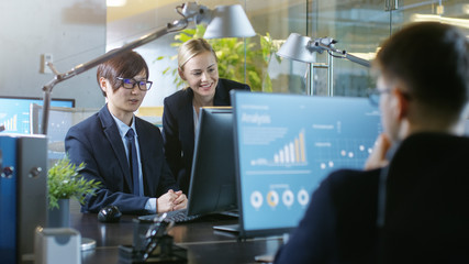 In the Office Businessman Sits at His Desk Talks with His Colleague, they're pointing at Computer Screen. Professional Businesspeople in Stylish Office.
