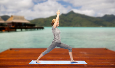 fitness, sport, people and healthy lifestyle concept - woman making yoga warrior pose on wooden pier over island beach and bungalow background