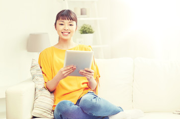 people, technology and leisure concept - happy young asian woman sitting on sofa with tablet pc computer at home