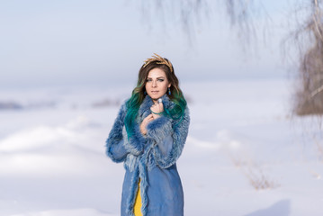 Beautiful girl in a blue fur coat in winter in nature. A snowy field. Blue hair. Wheat ears in hair. Hands on the collar of a fur coat.