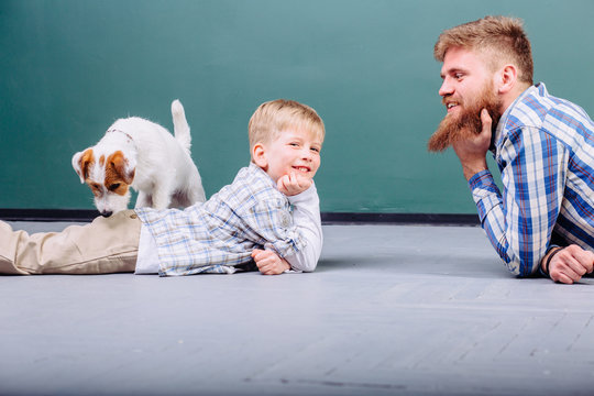 Happy Caucasian family having fun in their new home playing with puppy dog jack russell terrier. Hansome beard father and his preschooler son lying on the floor in front of each other. Side view