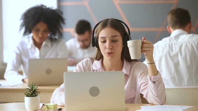 Funny happy young woman singing while listening to loud music in headphones sitting at office desk with laptop surrounded by diverse colleagues, lazy worker wasting time at workplace in co-working