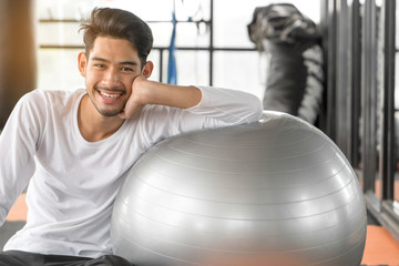 Fototapeta na wymiar Healthy workout. Young cheerful man sitting on the yoga mat and exercising with ball in the gym.