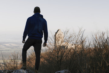 Adult man (hiker) standing on the rock and enjoying view with blurred castle on background. Silhouette of tourist man in forest at sunset.