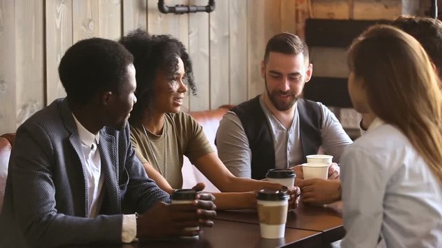 Multi-ethnic best friends enjoy drinking hot coffee at meeting in cozy coffeeshop, diverse young people chatting spending time in coffeehouse, multiracial fellows talking joking sitting at cafe table