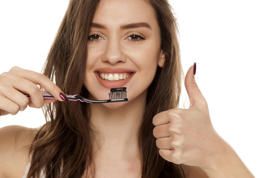 young woman holding a black tooth paste with active charcoal, and black tooth brush on white background, and showing thumbs up