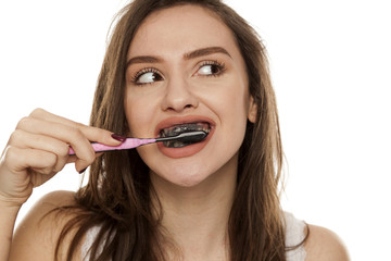 young woman brushing her teeth with a black tooth paste with active charcoal, and black tooth brush on white background - obrazy, fototapety, plakaty