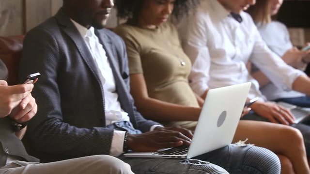 Multi-ethnic casual people waiting in queue ignoring each other immersed in phones apps and laptops, diverse young students sitting with devices together. People and technology concept. Close up view