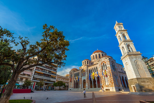 Stone Greek Orthodox Church on the town square with the city clock on the tower, among modern houses, summer, blue clear sky. Cathedral Church of St. Nicholas, Volos, Greece - April 2017.