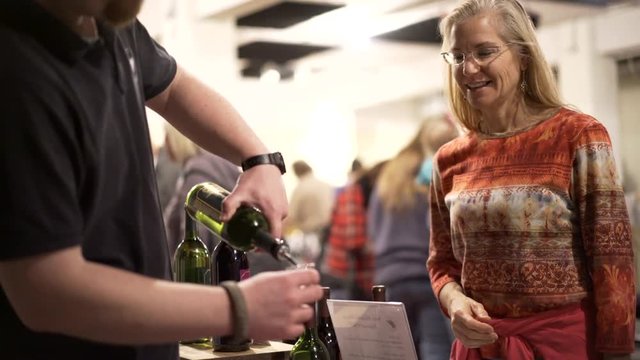 Beautiful, mature woman watches as wine sample is poured at wine and food festival indoors