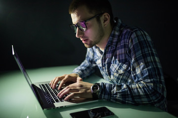 Concentrated young hacker using computer and stealing information in dark room in the night.