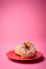 Two glazed donuts, one with colorful sprinkles, on a red plate with a pink background.