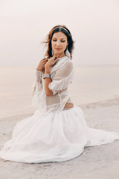 beautiful young stylish boho woman sitting on the beach at sunset
