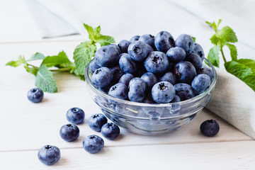 Ripe blueberries in a glass bowl and mint on a light table.