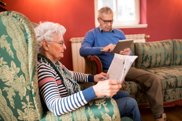 Senior couple relax at home while reading newspaper and using tablet